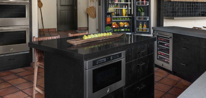 A kitchen with an opened JennAir Refrigerator filled with beverages and produce, a JennAir Undercounter Refrigerator and a JennAir Microwave built into an island. On the island is a tray of pears.